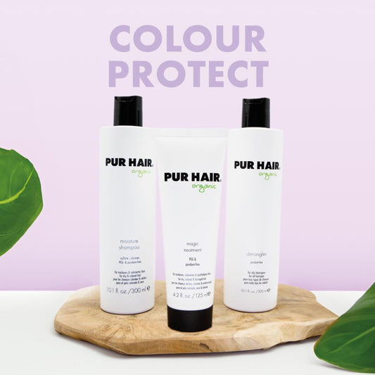 PUR HAIR Organic Colour Protect Set bestehend aus Moisture Shampoo, Magic Treatment und Detangler. Die drei weißen Flaschen und Tuben stehen dekorativ auf einem Holzbrett vor einem pastell-lila Hintergrund mit der Aufschrift ‚Colour Protect‘