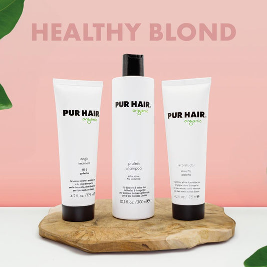 PUR HAIR Organic Blond Set bestehend aus Protein Shampoo, Reconstructor und Magic Treatment. Die drei weißen Flaschen und Tuben stehen dekorativ auf einem Holzbrett vor einem pastell-rosa Hintergrund mit der Aufschrift ‚Healthy Blond‘