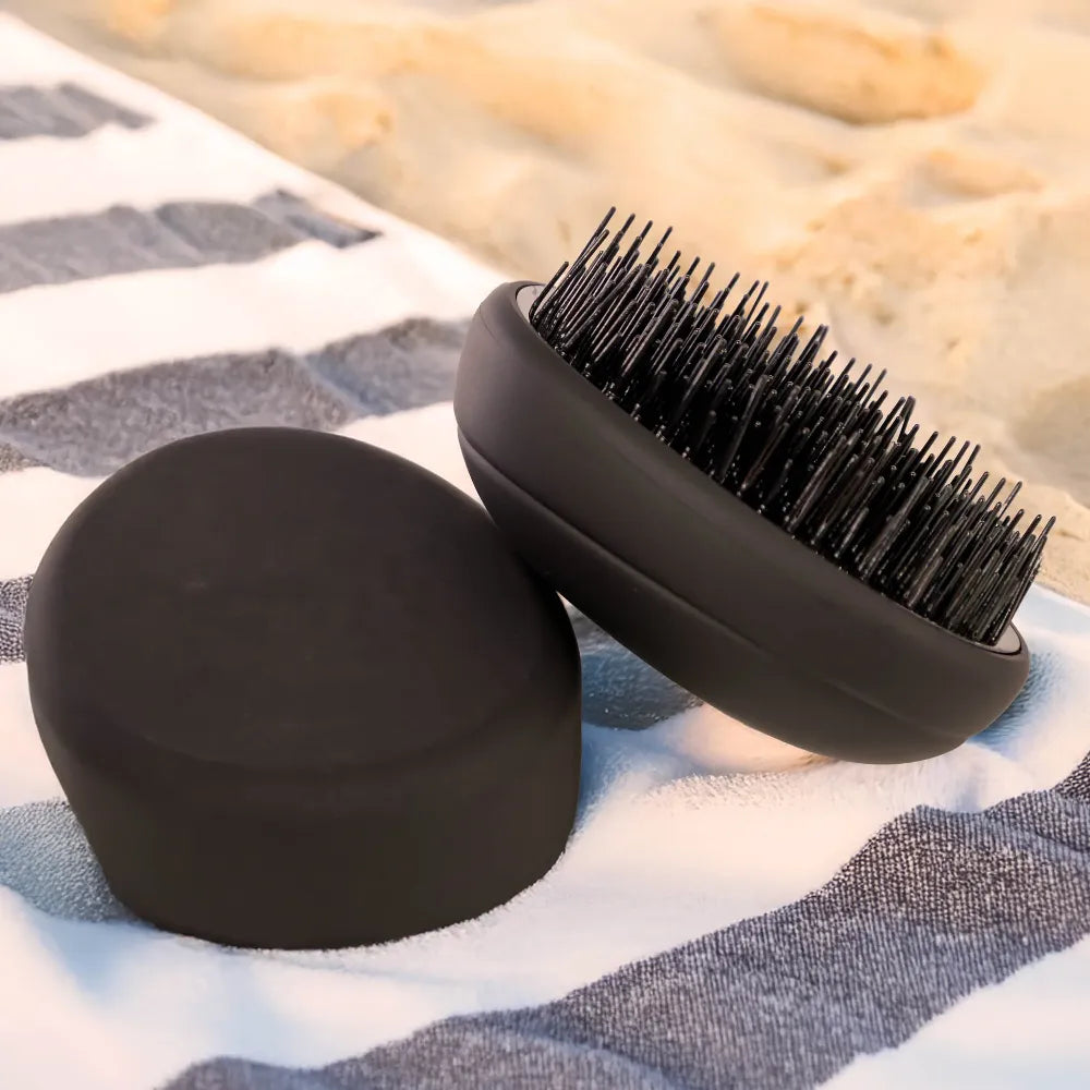 PUR HAIR Detangler Brush, in schwarz, liegt auf gestreiftem Handtuch, am Strand