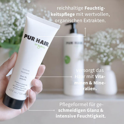 PUR HAIR Organic Moisture Treatment in 125 ml Tube wird in der Hand gehalten, im Hintergrund weiße Blüten und eine große Flasche; Text weist auf Feuchtigkeitspflege, Vitamine und Mineralien hin