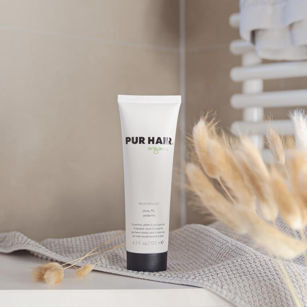 Weiße 125-ml-Tube PUR HAIR Organic Reconstructor mit schwarzem Deckel, aufgestellt neben Vase und Pflege-Accessoires