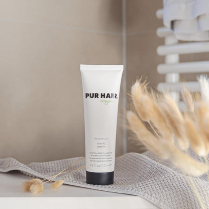Weiße 125-ml-Tube PUR HAIR Organic Reconstructor mit schwarzem Deckel, aufgestellt neben Vase und Pflege-Accessoires