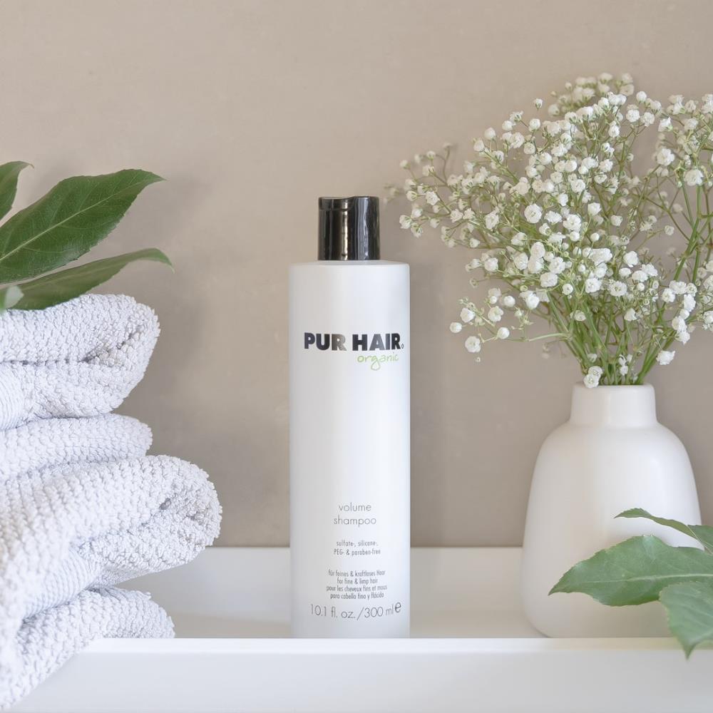 Weiße Flasche PUR HAIR Organic Volume Shampoo, 300 ml, neben grauen Handtüchern mit  Trockenblumen im Badezimmer platziert