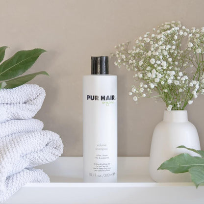 Weiße Flasche PUR HAIR Organic Volume Shampoo, 300 ml, neben grauen Handtüchern mit  Trockenblumen im Badezimmer platziert